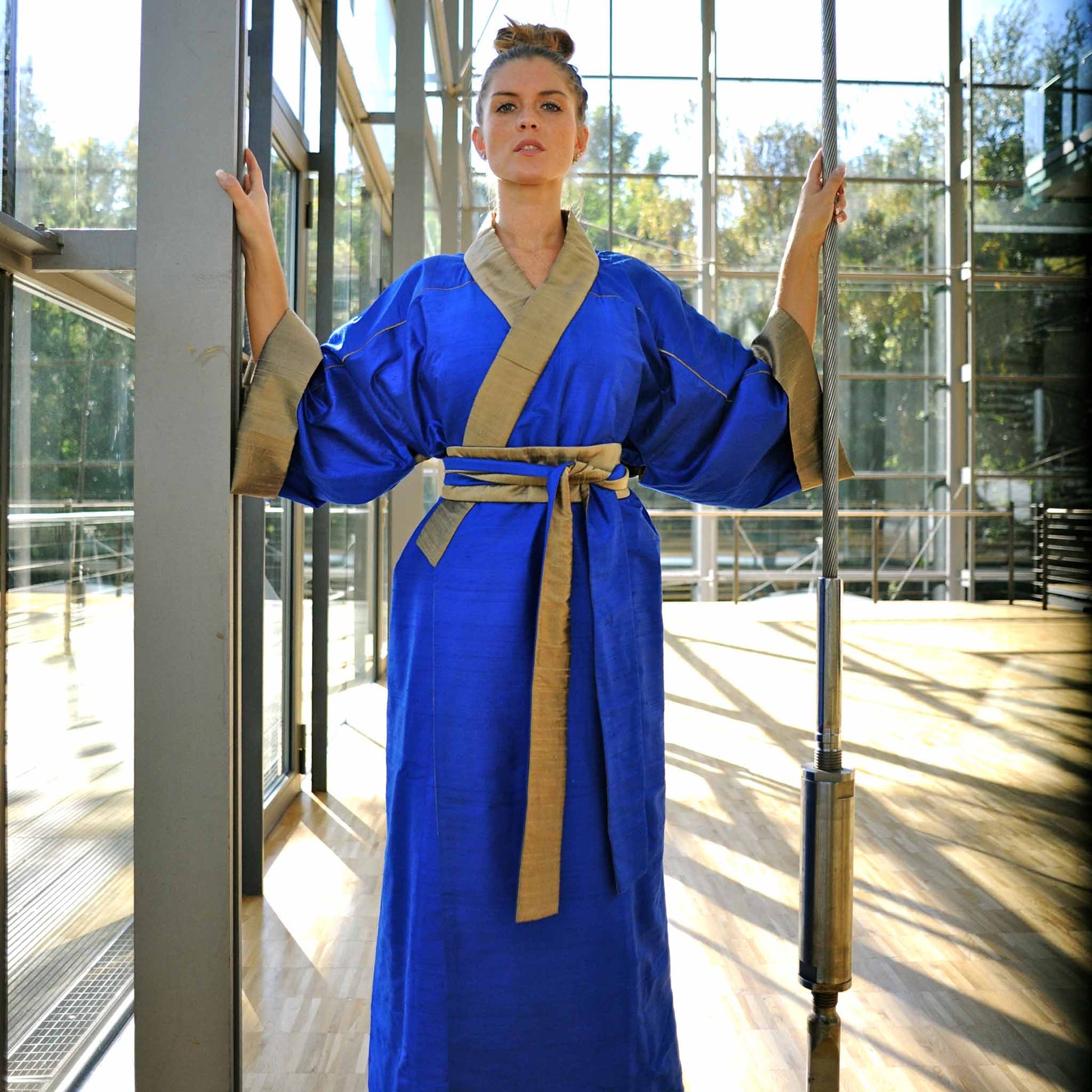 Eine junge Fraugeht steht frontal zur Kamera. Sie trägt einen SeidenKimono von Design Gianni Sarto, mit den Farben Blau und gold..