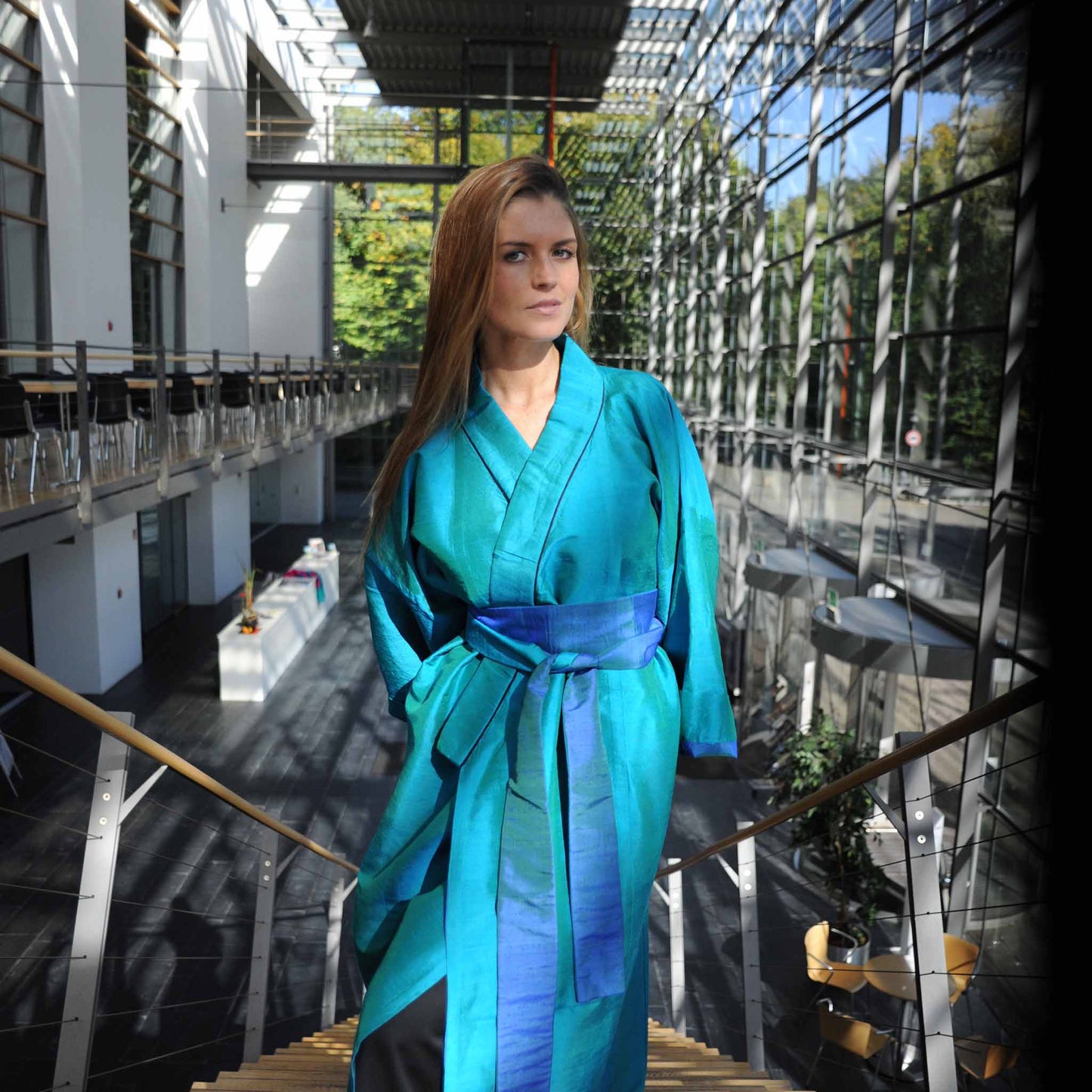 Eine junge Frau kommt dem Betrachter auf einer Treppe entgegen. Sie trägt einen Kimono von Design Gianni Sarto, mit den Farben Grün und Blau.